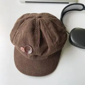 Braune Schiebermütze in Cord-Optik mit großem Knopf, flach neben Kopfhörern fotografiert