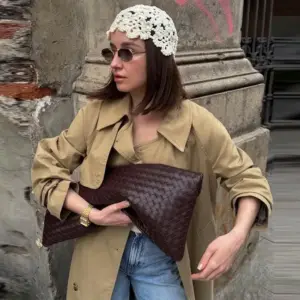 Frau mit cremefarbener gehäkelter Mütze mit Blumenmuster, Trenchcoat und Clutch