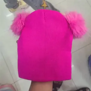 Pinkfarbene Strickmütze mit zwei flauschigen Bommeln, von einer Hand gehalten