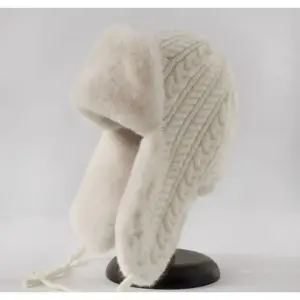 01-39 Cremefarbene Strickmütze mit Zopfmuster, flauschigen Ohrenklappen und herabhängenden Bändern auf dunklem Ständer