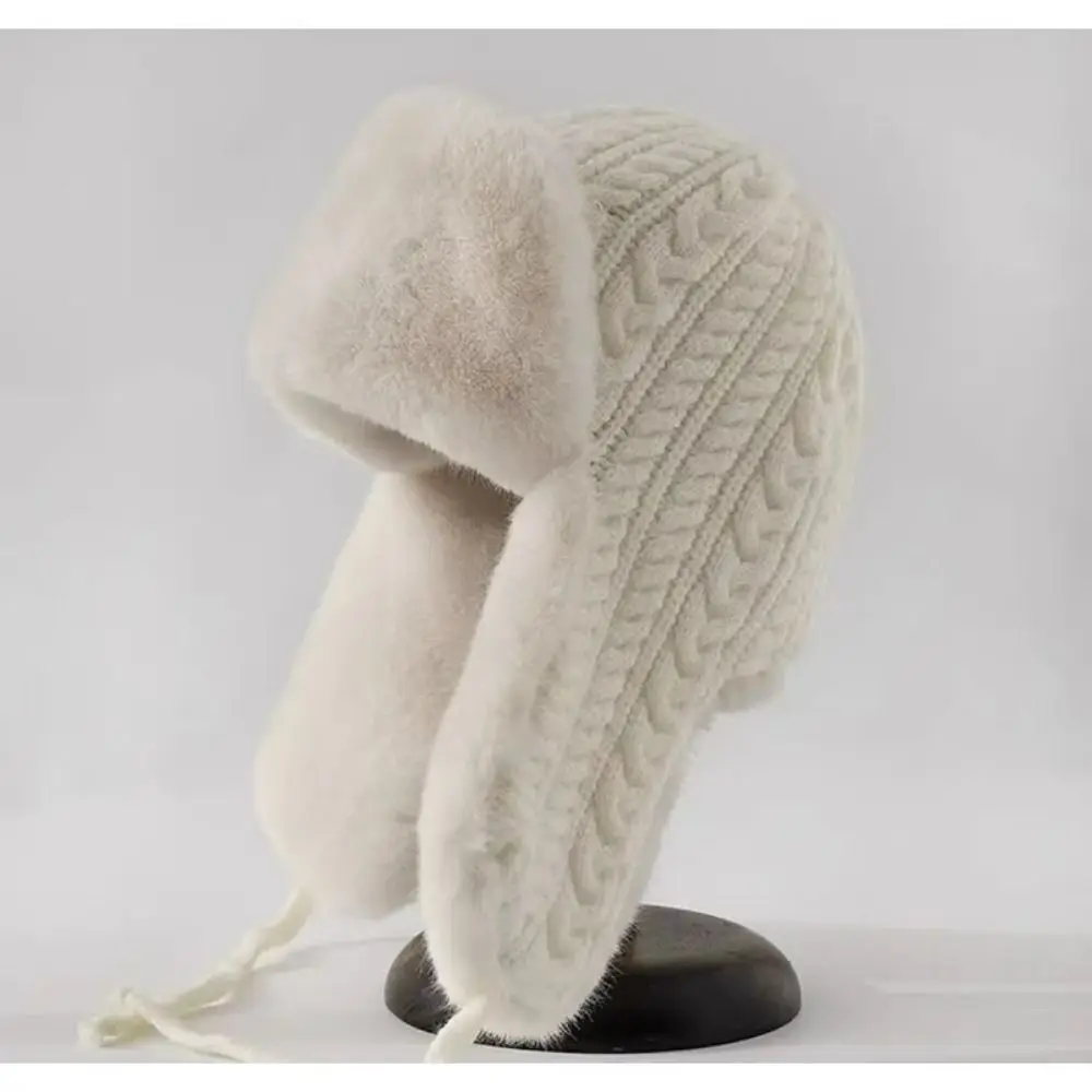Cremefarbene Strickmütze mit Zopfmuster, flauschigen Ohrenklappen und herabhängenden Bändern auf dunklem Ständer