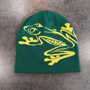 Grüne Strickmütze mit großem Frosch-Motiv in Kontrastfarbe, flach auf grauem Untergrund liegend