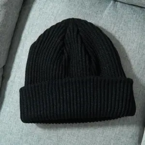 Schwarze grob gerippte Strickmütze mit Umschlag, flach auf einem grauen Sofa liegend